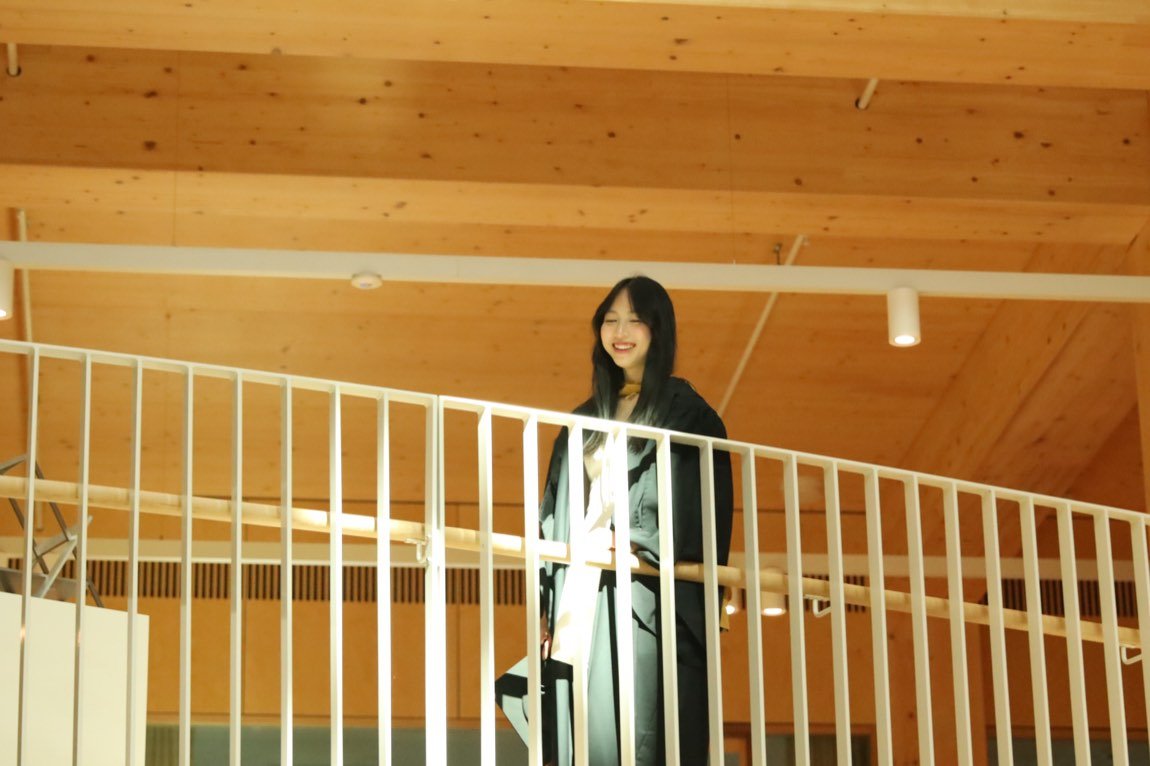 Graduate smiling from a balcony under a warm timber ceiling