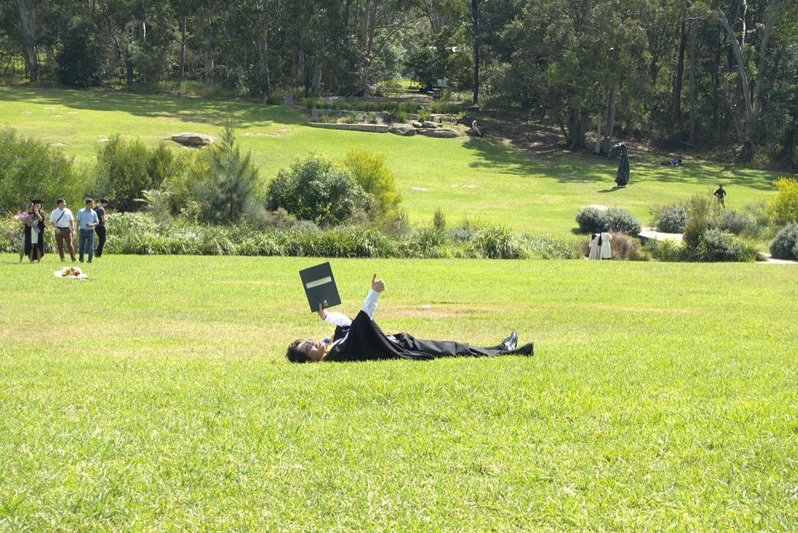 Graduate lying on the lawn holding his testamur with a thumbs up