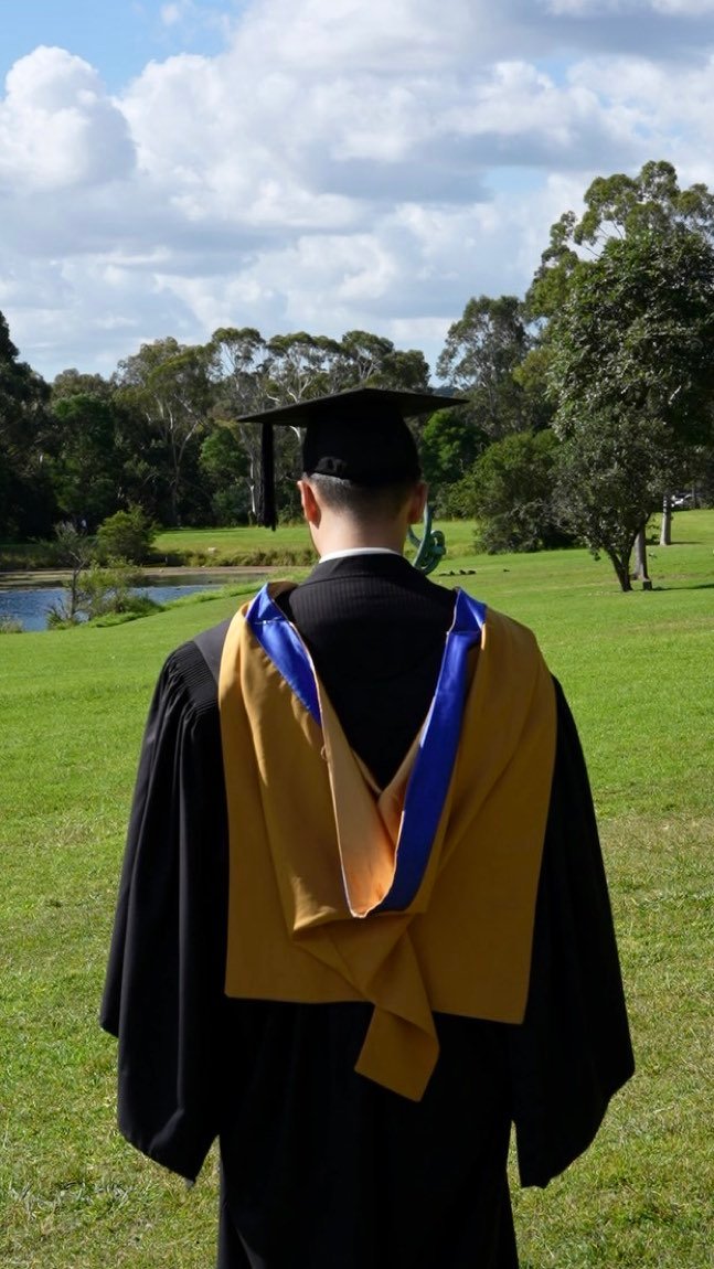 Graduate seen from behind showing the gold and blue hood by the lake