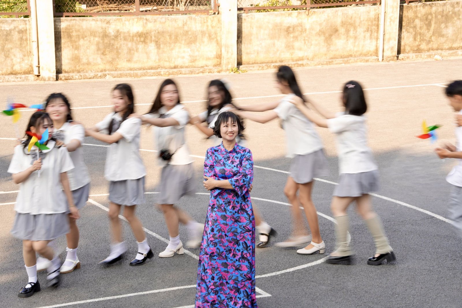 Teacher standing still while students in white uniforms spin around her in golden light